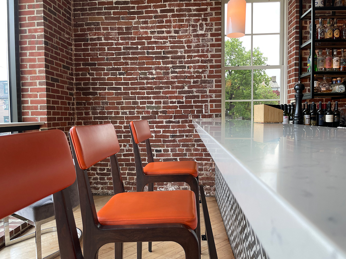 White marble top bar with dark wood bar stools that have burnt orange cushions. Brick Wall stright ahead with window letting light in.