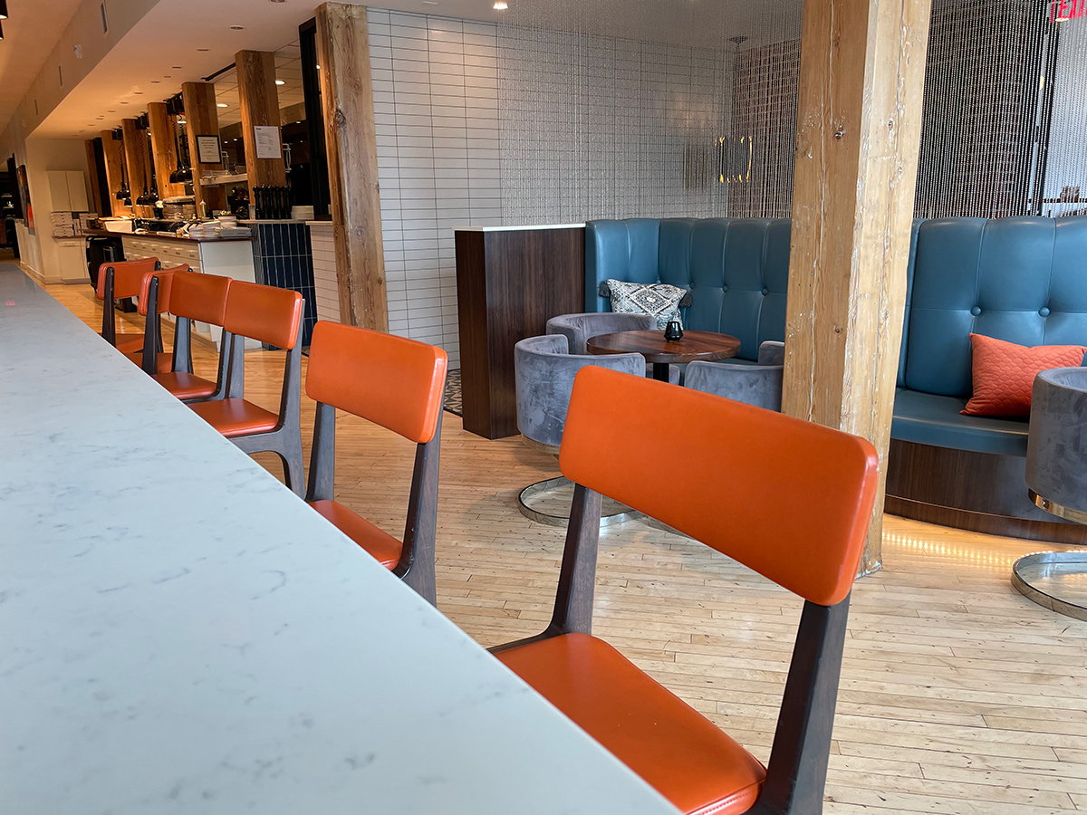 Photograph of interior of Cafe, White marble top bar in the foreground with Wooden bar chairs with burnt orange padding. In background there are booths with teal  cushions and C shaped grey suede table chairs. Overlooking a hallway with an open Kitchen featuring stainless steal shelves with plates and utensils.