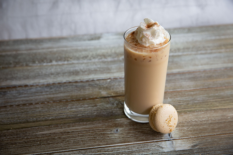 Iced Caramel Macchiato with a light creamy brown color, topped with fluffy whip and a Macaroon Cookie on the side. Set on a grey and whitewashed table and light background.