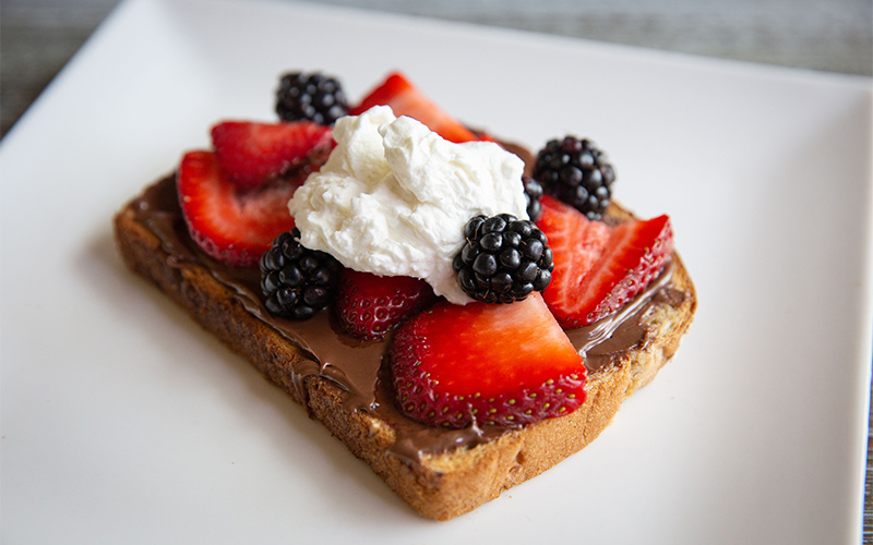 Piece of Toast topped with Nutella Hazelnut spread, strawberries, blackberries and Homemade whip sitting on a white plate.