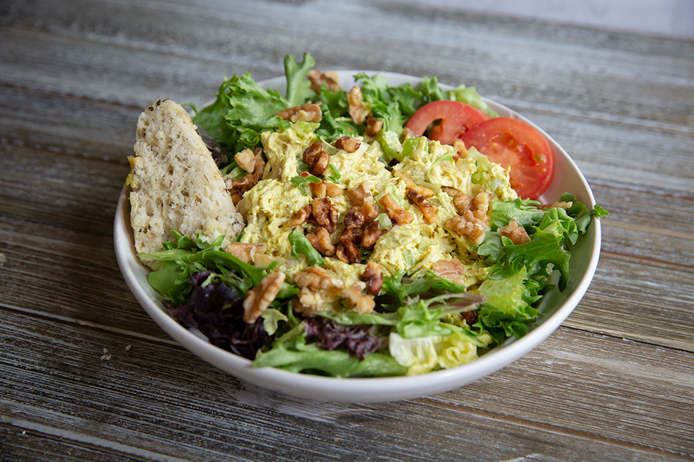 Large white serving bowl filled with mixed green and topped with a Curried chicken salad with grapes, onion , nuts and celery. With a side of a triangular soft scone.