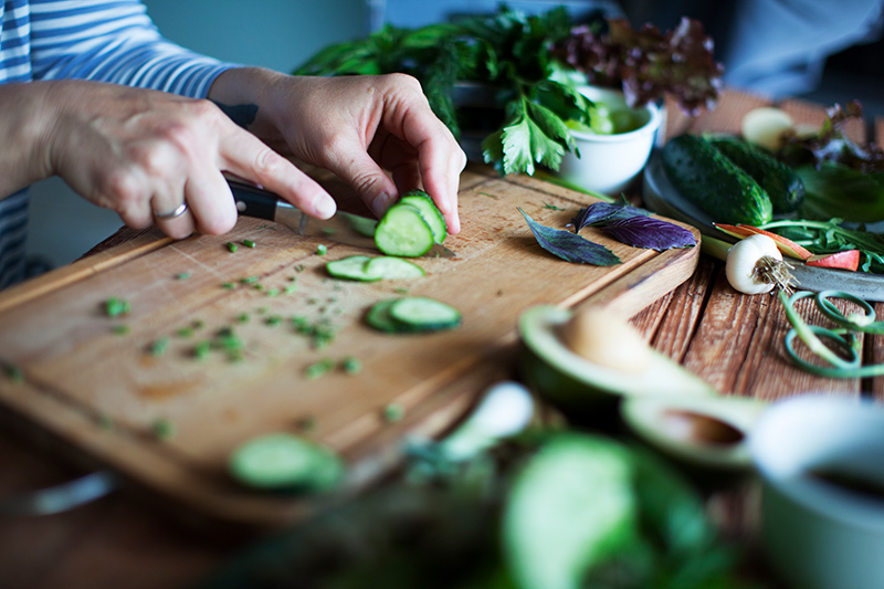 Woman's hands cutting cucumber on light brown cutting board, on a table with lots of fresh produce.