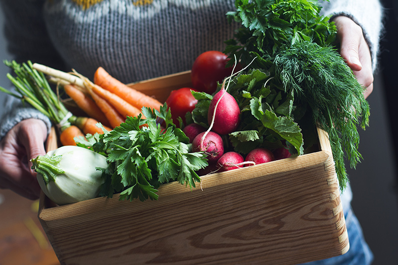 Woman's hands holding light brown solid wooden crate with lots of fresh produce.