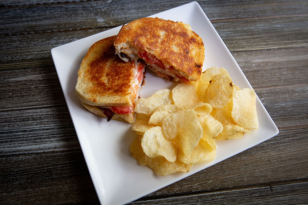 Top down image of grilled cheese sandwhich on white plate. Filled with Turkey, chedder, and asiago cheese, and bacon. It has kettle chips on the side.