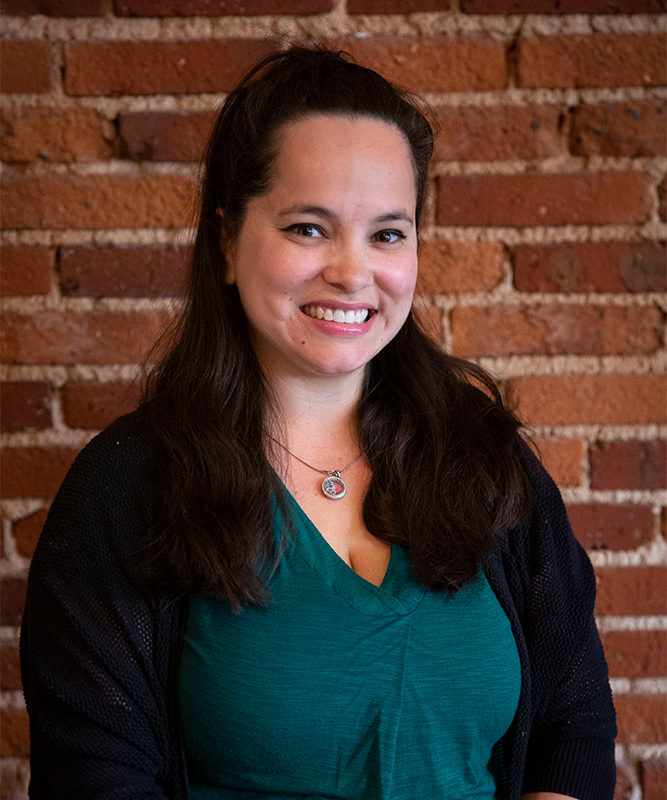 Woman in 30s with dark brown hair past shoulders and wide smile, wearing a teal shirt with black jacket, standing in front of brick wall.