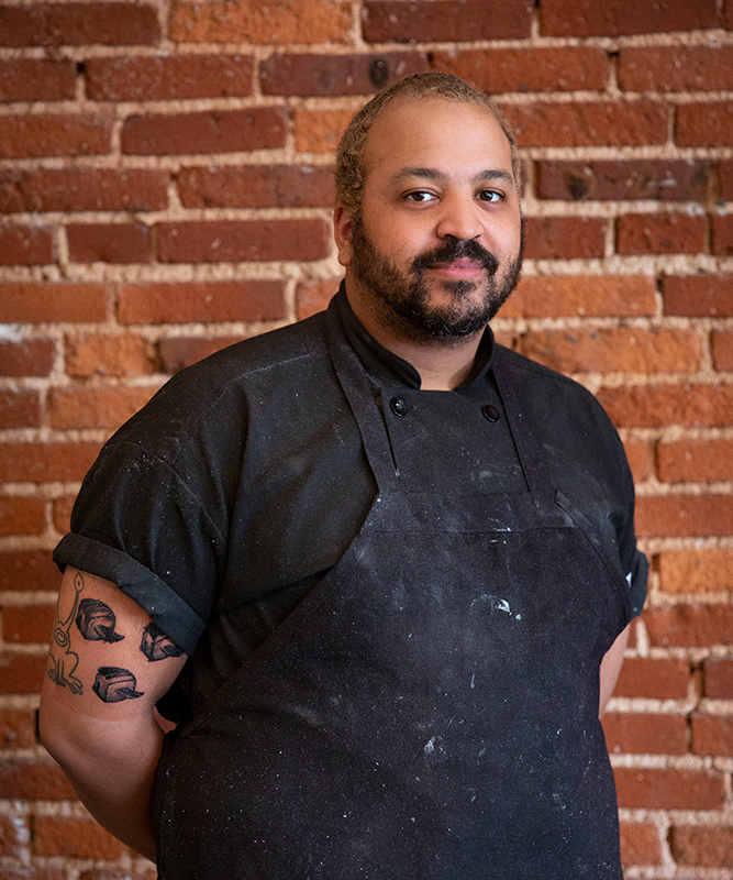 Man in 30s with very short dark brown and blonde hair and dark beard, wearing black chefs apron and black shirt, standing in front of brick wall.