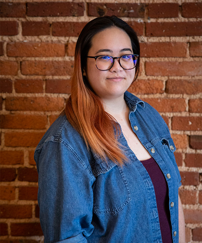 Woman in 20s with dark brown hair and Red/brown highlights past shoulders and glasses, wearing a maroon shirt and jean jacket, standing in front of brick wall.